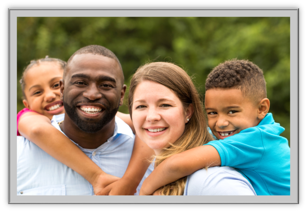 A smiling family poses outdoors, with two children playfully embracing their parents' shoulders. The background is a vibrant, blurred green.