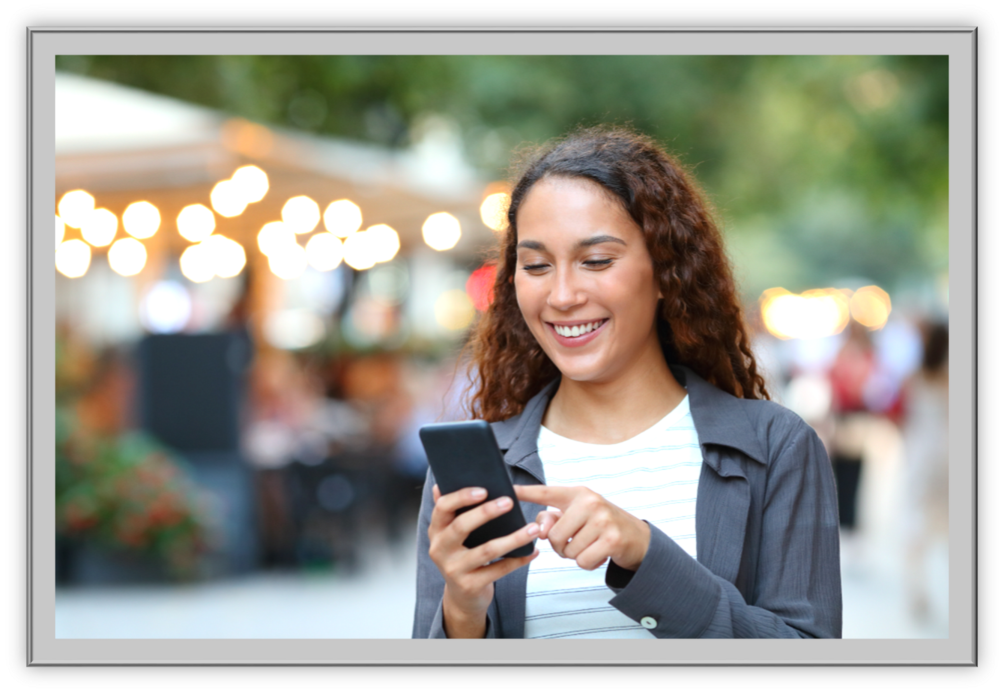 Smiling woman with curly hair uses a smartphone outdoors. Blurred festive lights and greenery create a cheerful, lively atmosphere in the background.