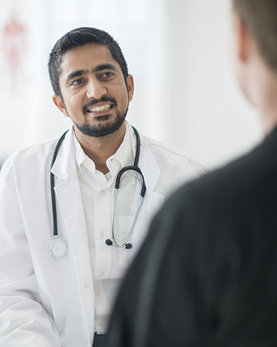 A doctor in a white coat and stethoscope smiles while talking to a patient. The setting is a bright, clinical office. The tone is friendly and reassuring.