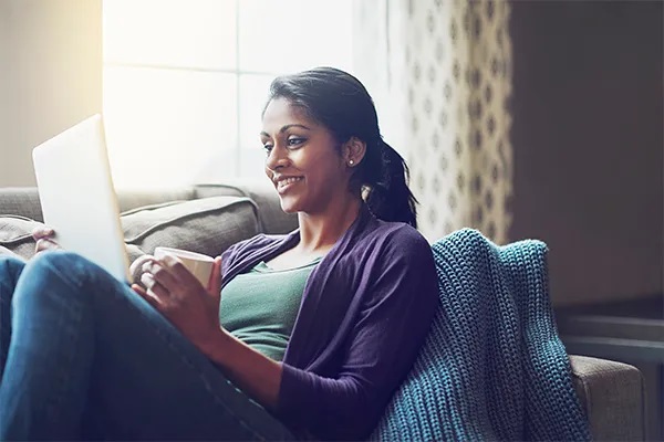 A woman relaxes on a couch, smiling at a laptop screen with a cup in hand. She wears a casual outfit, and a knit blanket is draped beside her.