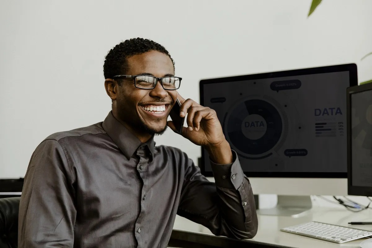 Smiling man in glasses and dark shirt, talking on phone. He's seated at a desk with a computer displaying data visualization, suggesting a productive work environment.