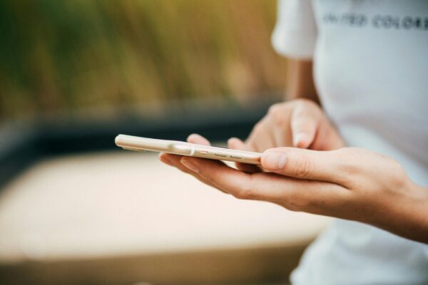 A person wearing a white shirt is holding a smartphone, focusing intently on the screen. The background is softly blurred, suggesting an outdoor setting.