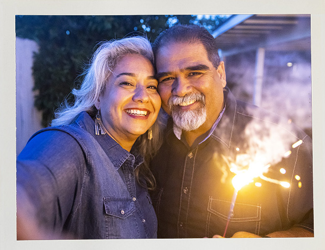 A joyful couple, dressed in blue, smiles warmly while holding a lit sparkler outdoors at dusk. The scene conveys happiness and celebration.