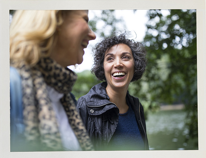 Two women laughing together outdoors by a lake, surrounded by trees. The atmosphere is joyful and relaxed, with soft, natural lighting.