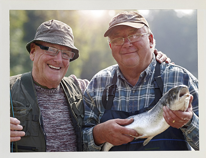 Two smiling elderly men in fishing gear stand together outdoors. One holds a fish, showcasing a sense of achievement and camaraderie in nature.