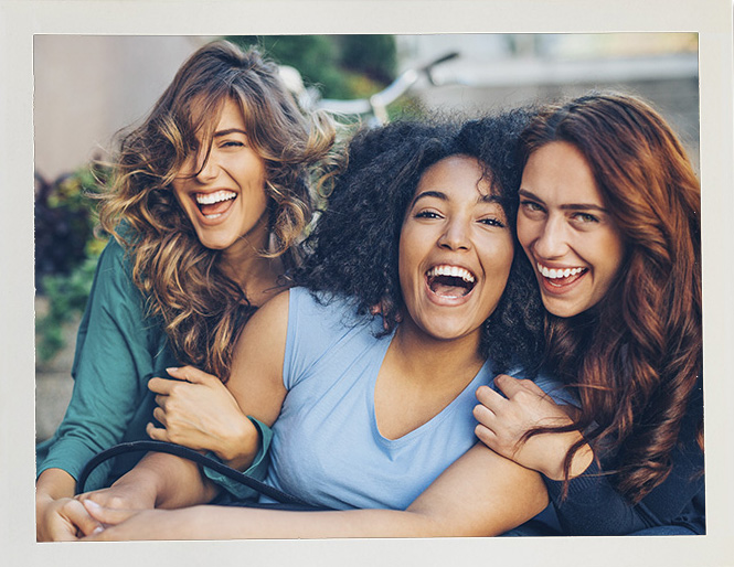 Three women laughing and embracing outdoors, exuding joy and friendship. They are sitting closely together, capturing a moment of happiness.