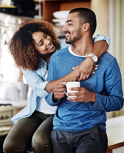 A smiling woman embraces a man holding a coffee cup in a cozy kitchen setting, both showing warmth and affection. Shelves with dishes are in the background.