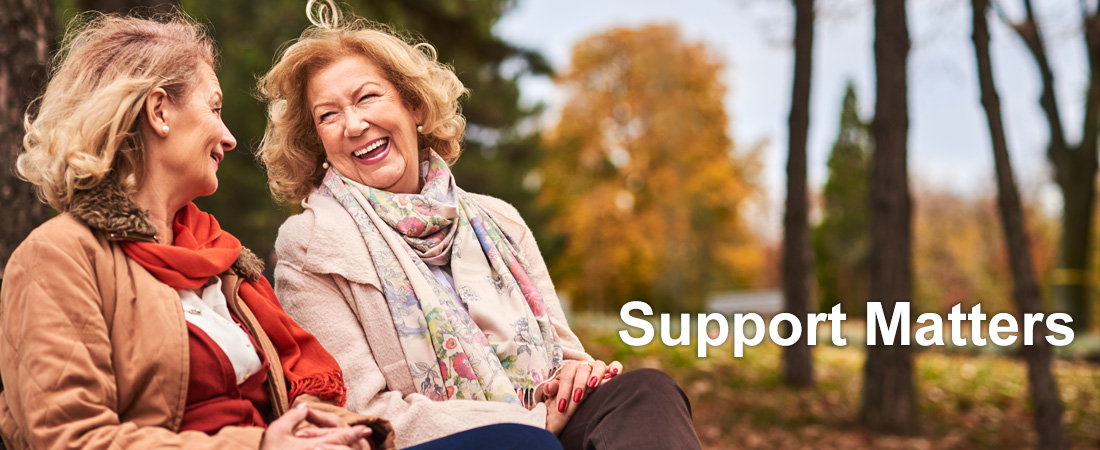 Two older women sit on a park bench, smiling and chatting warmly. Autumn leaves and trees surround them. Text beside them reads, Support Matters.